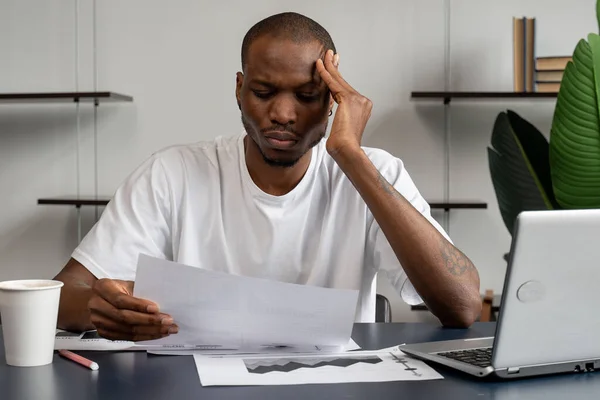 Un joven hombre de negocios negro parece desconcertado, tiene problemas, está sentado en una pose de pensamiento profundo, mirando los papeles — Foto de Stock