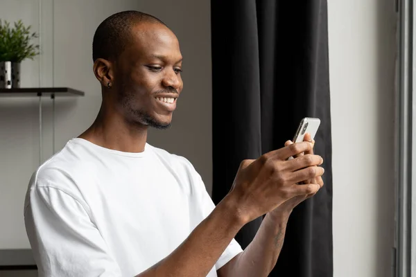 Joven negro chateando con amigos en una red social, escribiendo mensajes, leyendo un buen correo electrónico mientras está de pie en la ventana — Foto de Stock