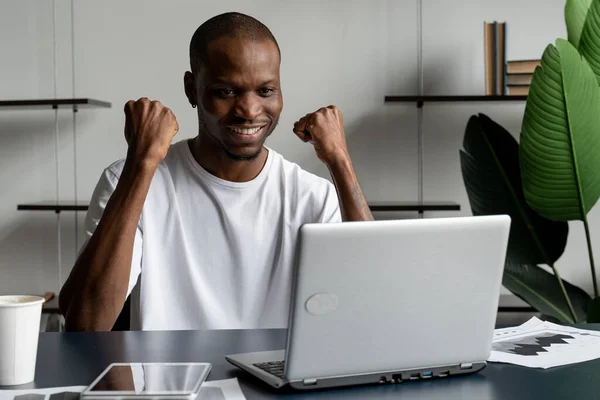 Feliz empresario emocionado celebra su éxito. El ganador, un hombre negro en la oficina leyendo las noticias en un portátil — Foto de Stock
