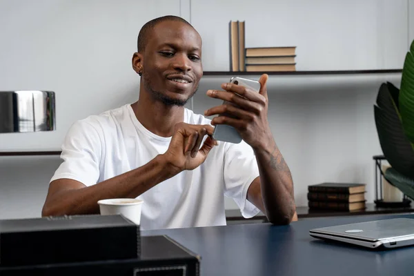 El hombre negro usa un teléfono inteligente mientras está sentado en un escritorio en la oficina. Un joven feliz comunicándose con colegas y clientes a través de Internet. — Foto de Stock