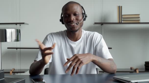 Lächelnder schwarzer Mann mit Headset, der an einem Laptop arbeitet und in die Kamera schaut, während er in der Kundenserviceabteilung arbeitet — Stockvideo