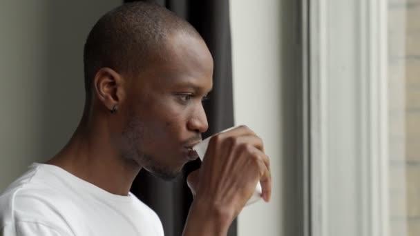 Guapo joven negro sostiene una taza de bebida caliente mientras mira por la ventana — Vídeos de Stock