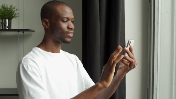 Młody czarny facet rozmawiający z przyjaciółmi w sieci społecznościowej, wpisujący wiadomości, czytający miłego maila stojąc przy oknie — Wideo stockowe