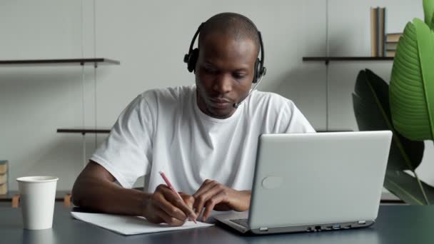 Skupiony czarny biznesmen noszący słuchawki, piszący notatki w notatniku, oglądający webinarium z kursu wideo — Wideo stockowe