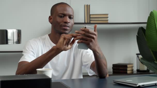 Schwarzer Mann benutzt Smartphone, während er an einem Schreibtisch im Büro sitzt. Ein glücklicher junger Mann, der mit Kollegen und Kunden über das Internet kommuniziert. — Stockvideo
