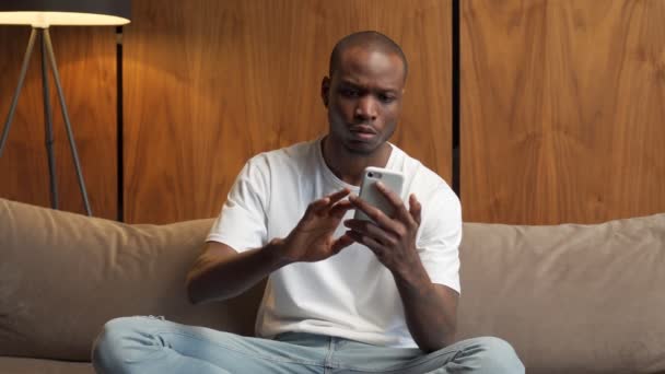 Hombre negro alegre usando un teléfono inteligente está sentado en casa en el sofá, celebrando una victoria, ganando una lotería en línea, regocijándose en la victoria — Vídeos de Stock