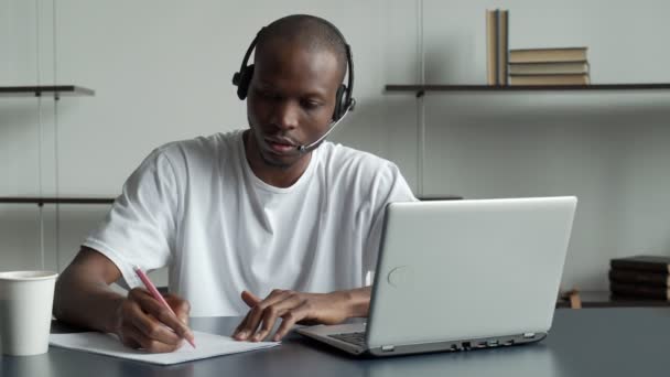 Estudiante negro con auriculares, estudia en línea, realiza ejercicios con una computadora portátil, ve videos, escribe en un cuaderno — Vídeo de stock