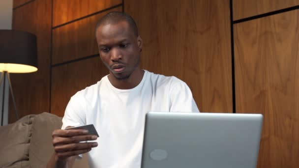Homem negro jovem que possui tecnologia moderna, um cartão de banco de plástico, sentado em um sofá confortável, fazendo compras usando um laptop — Vídeo de Stock