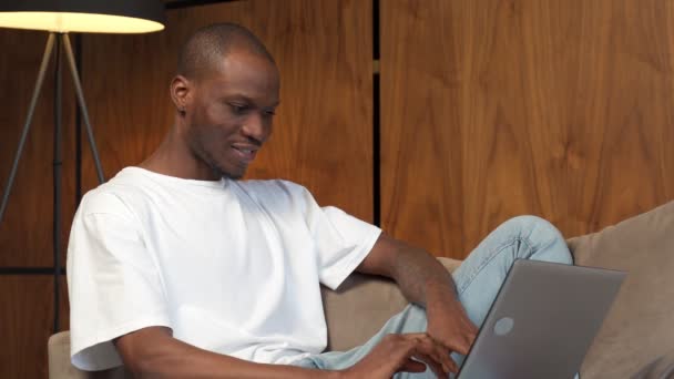 Junger glücklicher schwarzer Mann kommuniziert online auf einem Laptop. Zufälliger Kerl sitzt auf der Couch im Wohnzimmer — Stockvideo