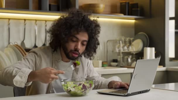 Homem árabe come salada trabalhando em laptop à mesa na cozinha — Vídeo de Stock