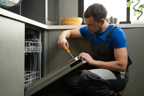 Repairman repairs dishwasher with screwdriver in kitchen