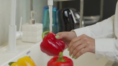 Genç bir kadın mutfakta musluk suyunun altında kırmızı biber yıkıyor.