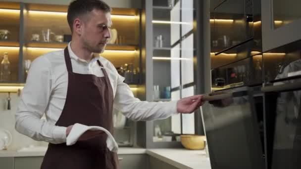 Чоловік в фартусі виймає свіжу пекарню з печі на кухні — стокове відео