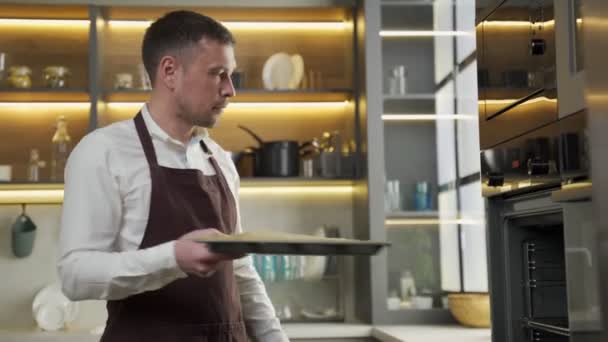 Mężczyzna szef kuchni w fartuchu wkłada surowe ciasteczka do piekarnika do pieczenia — Wideo stockowe