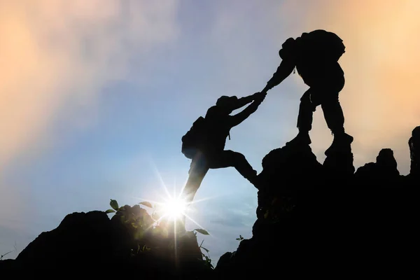 Silhouette of two people climbing a mountain in a trekking activity at sunset, teamwork assistance concept