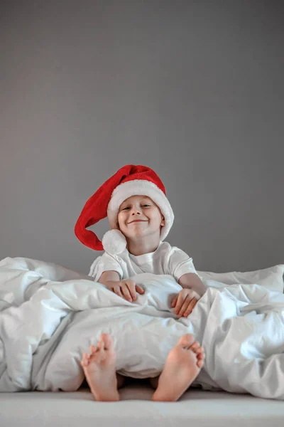 Lustige süße Junge sitzt auf dem Bett schlürft in einem Weihnachtsmann Hut. Frohe Feiertage. Konzept für den Weihnachtsmorgen — Stockfoto