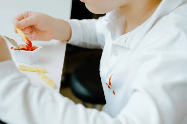 Fechar as manchas sujas de ketchup em roupas de criança branca. Menino comendo batatas fritas batatas fritas e molho de tomate na mesa em um restaurante de fast food. Crianças comem alimentos gordurosos não saudáveis. — Fotografia de Stock