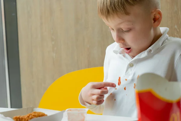 Çocuk bir fast food restoranının masasındaki elbiselerindeki ketçap lekesini işaret ediyor. Çocuklar sağlıksız yağlı yiyecekler yerler. Kıyafetlerdeki leke temizleme kavramı.