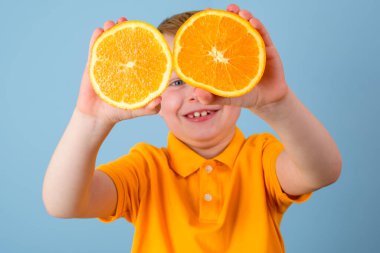 Olumlu duygular. Gözlerinde turuncu olan süpriz bir çocuk. Elleriyle portakalın iki yarısını tutan neşeli bir çocuk. Mavi arka planda izole edilmiş. Sarı tişörtlü çocuk. Yüksek kalite fotoğraf