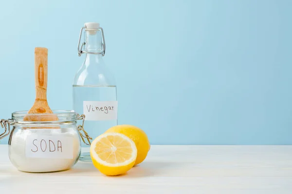 Open jar of baking soda with a wooden spoon on top, vinegar, cut lemon, on a blue background. The concept of organic removing stains on clothes.