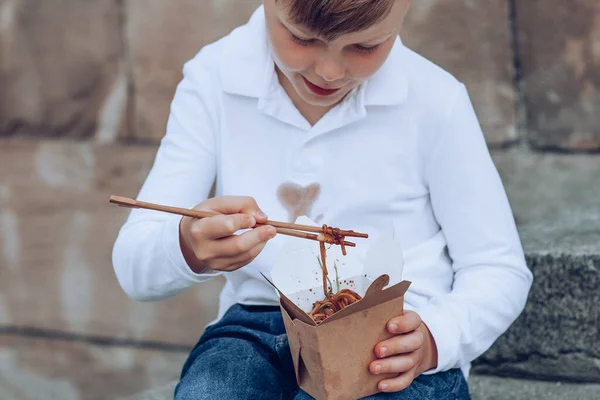The child eating on the go soy sauce pan-fried noodles chopsticks. Dirty soy sauce stains on a white t-shirt. Takeaway food. outdoors. High quality photo