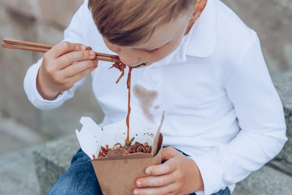 The child eating on the go soy sauce pan-fried noodles chopsticks. Dirty soy sauce stains on a white t-shirt. Takeaway food. outdoors. High quality photo