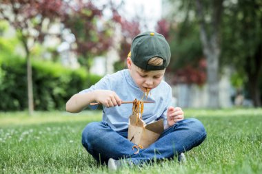 Tişörtte kirli soya sosu lekesi var. Elinde soya soslu erişteyle birlikte yemek çubuğu tutan çocuk otların üzerinde oturuyordu.. 