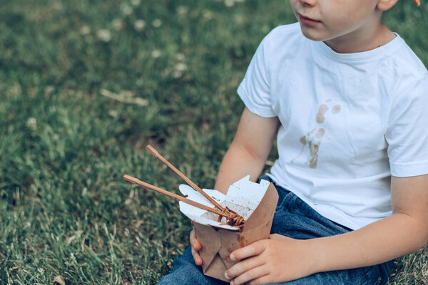 The boy holding takeaway food sitting on the grass. Dirty sauce stains on a white t-shirt. outdoors. High quality photo
