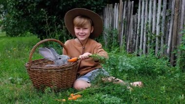 A rabbit eating a carrot, looking out of the basket. A boy in a hat feeding an animal while sitting on the grass. outdoor. High quality photo