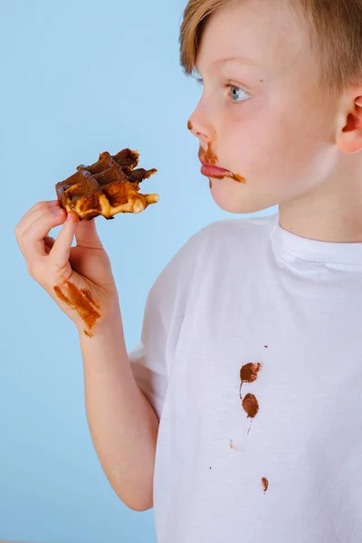 Tシャツに汚れたチョコレートの汚れ 青い背景にチョコレートの甘いソースでベルギーのワッフルを食べる少年 高品質の写真 — ストック写真