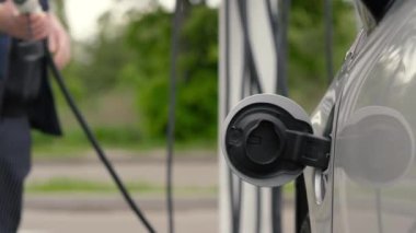 Tanınmayan adam EV vagonuna güç bağlantısı yerleştiriyor. Erkek el, elektrikli araca şarj kablosu takıyor ve pilleri şarj ediyor. Otoparktaki EV istasyonu