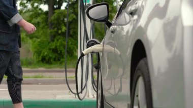 Tanımlanamayan sakallı adam elektrik bağlantısını koparıyor. Erkek el, şarj kablosunu elektro arabaya bağlar, pilleri şarj eder. EV istasyonu otoparkı yeşillikler arka plan