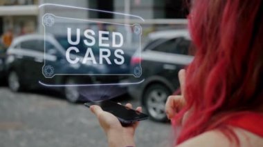 Unrecognizable redhead woman standing on street interacts HUD hologram with text Used cars. Lady in red clothes uses technology of the future mobile screen