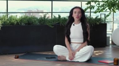 Woman is talking, sits cross-legged on karemat on terrace with greenery, participating in video conference, remotely negotiating with clients. Video tutorial for work or study