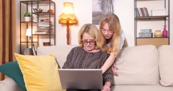 Oldalsó nézet a régi kaukázusi nagymama és unokája használja laptop kényelmes otthon. Nő unoka tanítja az idős nagymama használni laptop 4k — Stock videók