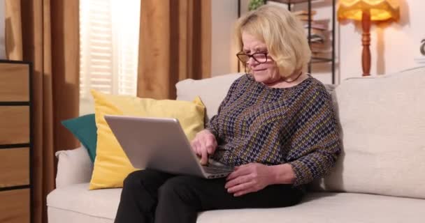 Plný pohled na šedovlasou starší ženu sedící doma na pohovce, používající přenosný počítač — Stock video