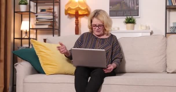 Πλήρης άποψη της γκριζομάλλης ηλικιωμένης γυναίκας που κάθεται στον καναπέ στο σπίτι, χρησιμοποιώντας φορητό υπολογιστή — Αρχείο Βίντεο