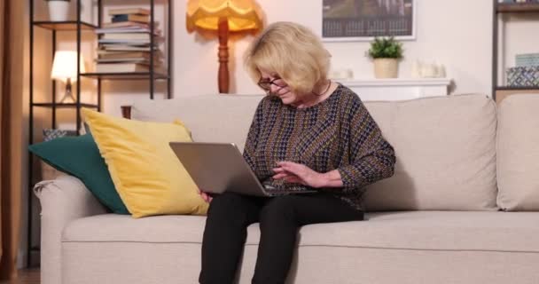 Vista completa della donna anziana dai capelli grigi seduta sul divano a casa, utilizzando il computer portatile — Video Stock