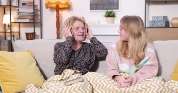 Abuela escuchando en los auriculares música favorita de la nieta, integración familiar. Chica feliz escuchando música en la tableta con su abuela en casa — Vídeos de Stock