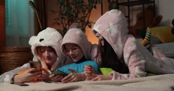 Trzy szczęśliwe dziewczyny w piżamie spędzają razem czas w domu, patrząc na telefon w pokoju badań.. — Wideo stockowe