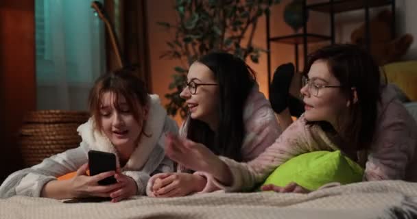 Tři šťastné dívky v pyžamu tráví spolu čas doma, dívají se na telefon v badroomu. — Stock video