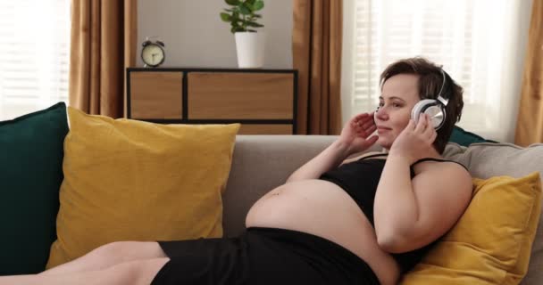 Glad gravid kvinna lyssnar musik, tar av hörlurar och håller nära magen — Stockvideo