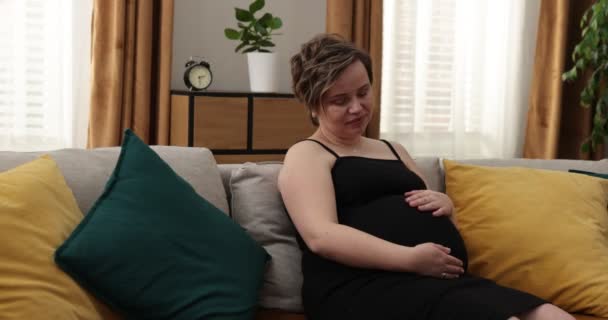 Glückliche Schwangere, die zu Hause ihren Bauch berührt. Schwangere liegt auf Sofa und streichelt ihren Bauch — Stockvideo