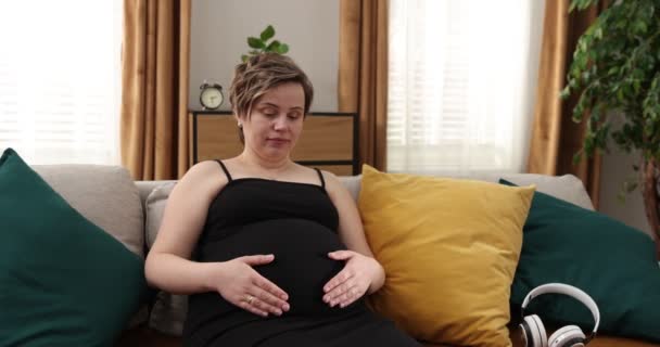 Gravid kvinna med hörlurar på magen. Kaukasiska gravid kvinna i sängen med hörlurar på bara magen och smeka den medan barnet lyssnar på musik — Stockvideo