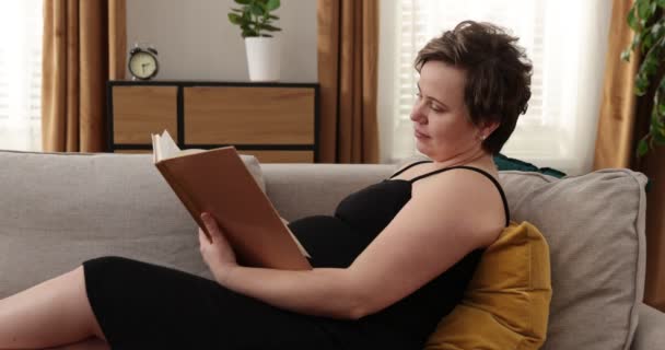 Ευτυχισμένη έγκυος καυκάσια γυναίκα διαβάζει βιβλίο στο σπίτι. Έγκυος αναπαύεται στο υπνοδωμάτιο — Αρχείο Βίντεο