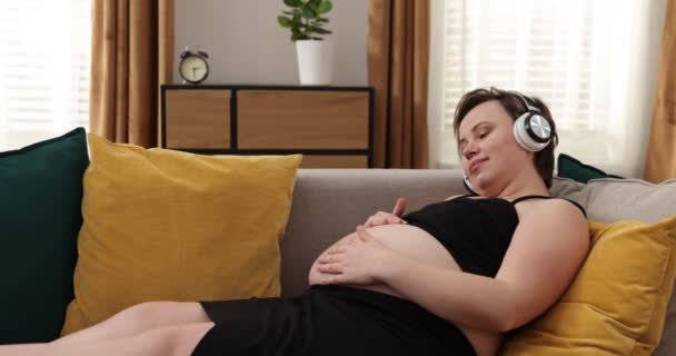 Těhotná žena drží sluchátka na břiše. Kavkazská těhotná žena v posteli drží sluchátka na holém břiše a hladí ji, zatímco dítě poslouchá hudbu — Stock video