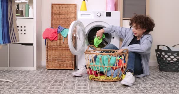 Genç çocuk kot elbiseli bir çamaşır makinesinin önünde oturuyor. Çamaşır makinesini kirli çamaşırlarla dolduruyor. Çamaşırhanede ev işi. — Stok video