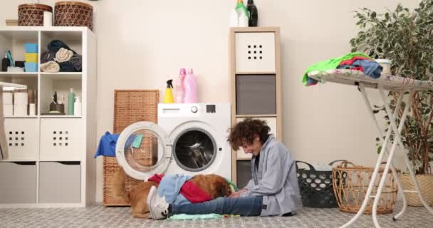 Молодий хлопчик сидить перед пральною машиною в одязі Джинсів. Він заряджає пральну машину брудною праскою. домашнє завдання в пральні — стокове відео