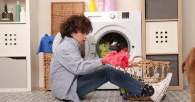 Kot pantolon ve gömlek giymiş gülümseyen genç bir çocuk bir çamaşır makinesinin önünde oturur. Kirli çamaşırları çamaşır makinesine yüklüyor. Parlak ve havadar bir çamaşır odası.