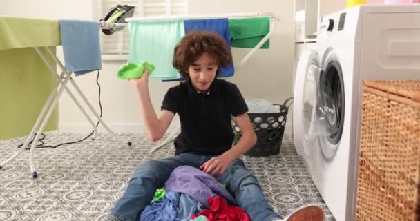 Kot pantolon ve gömlek giymiş gülümseyen genç bir çocuk bir çamaşır makinesinin önünde oturur. Kirli çamaşırları çamaşır makinesine yüklüyor. Parlak ve havadar bir çamaşır odası. — Stok video